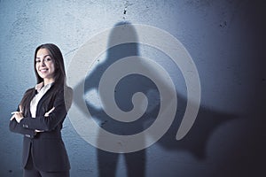 Attractive young european businesswoman with folded arms standing on concrete wall background with superhero cape shadow. Success