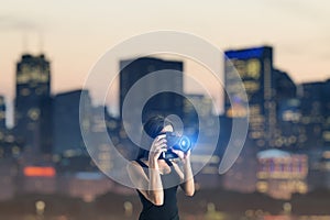 Attractive young european woman using glowing camera on blurry city skyscrapers background. Lifestyle, photographer and success