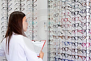Attractive young female doctor in ophthalmology clinic. Doctor ophthalmologist is standing near shelves with different eyeglasses