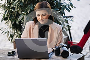 Attractive young female freelancer working on laptop on terrace in cafe. Woman photographer retouch photos on open air