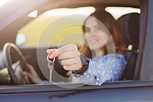 Attractive young female holds car keys, being happy owner of new automobile, blurred background. Lovely woman sells vehicle, adver