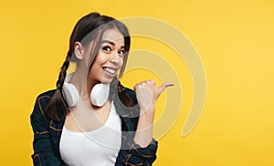 Attractive young female points on right, wearing casual shirt and white headphones, shows free space against yellow background