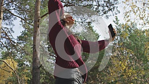 Attractive young female in red knitted jumper smiling happily and gesticulating while taking photos in sunlit autumn