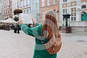Attractive young female tourist is exploring city. Redhead 30s woman with backpack holding a paper map on city street