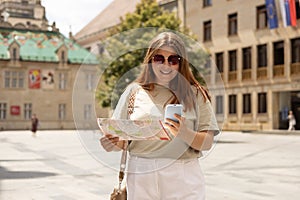 Attractive young female tourist is exploring city. Redhead 30s woman with backpack holding a paper map and phone on city