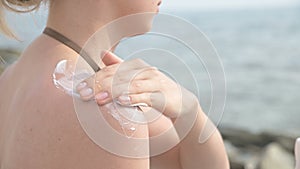 An attractive young girl applying sunscreen on her shoulder while sitting on the seashore on a sunny summer day. Summer