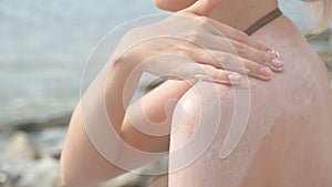 An attractive young girl applying sunscreen on her shoulder while sitting on the seashore on a sunny summer day. Summer