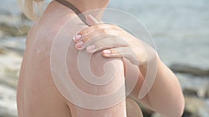 An attractive young girl applying sunscreen on her shoulder while sitting on the seashore on a sunny summer day. Summer