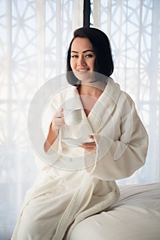 Attractive young girl in bathrobe holding a cup, looking at window and smiling while sitting on a bed near the big