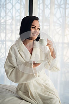 Attractive young girl in bathrobe holding a cup, looking at window and smiling while sitting on a bed near the big