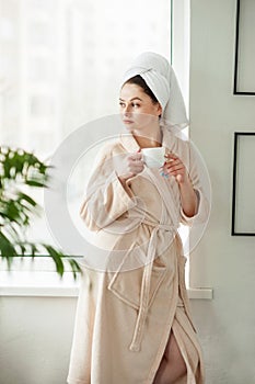 Attractive young girl in bathrobe and with a towel on her head is holding a cup, sitting on a window sill in the bathroom