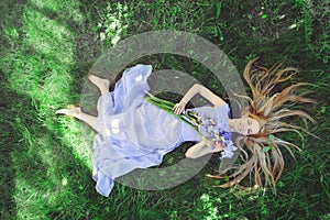 Attractive young girl with blonde hair and natural make-up smelling blue purple iris flowers lying on grass outdoors, tendern