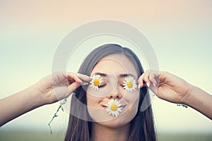 Attractive young girl having fun on summer field, holding daisies over her eyes and smiling. Nature, environment