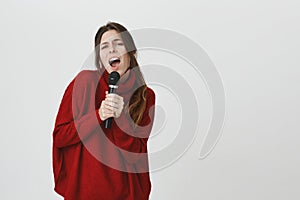 Attractive young girl in red winter sweater with long brown hair holding microphone with both hands and singing out loud