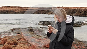 An attractive young girl stands on a rock on the seashore and looks at her phone. A hipster girl stands on the rocks