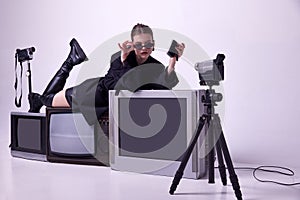 Attractive young girl in stylish black jacket, boots and sunglases lying on retro tv sets and recording video with