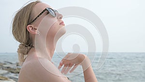 An attractive young girl in sunglasses applying sunscreen on her neck while sitting on the seashore on a sunny summer