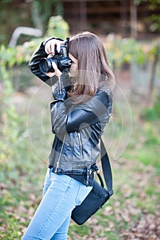 Attractive young girl taking pictures outdoors. Cute teenage girl in blue jeans and black leather jacket taking photos in park