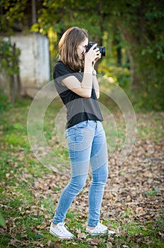 Attractive young girl taking pictures outdoors. Cute teenage girl in blue jeans and black t-shirt taking photos in autumnal park.