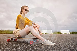 Attractive young girl with a tattoo on her arm in sunglasses and shorts sits on her longboard in a suburban parking on a