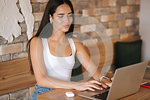 Attractive young girl use wirreless headphones and workin on her laptop. Woman in white t-shirt and denim. Student study