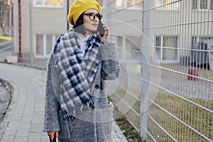 An attractive young girl wearing sunglasses in a coat walking down the street and talking on the phone and smiles