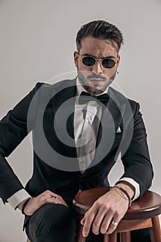 Attractive young groom in tuxedo with glasses holding elbow on wooden chair