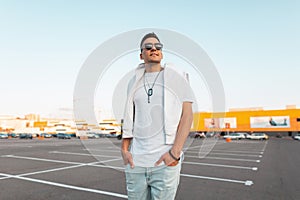 Attractive young happy man with a cute smile in fashionable summer clothes in stylish black sunglasses stands in the city parking