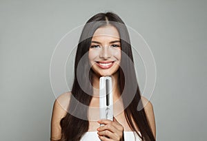 Attractive young healthy brunette woman holding hair iron and straightening her long smooth shiny hair on white background.