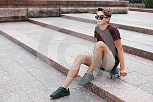 Attractive young hipster man in dark sunglasses in summer fashionable clothes in sneakers sits on a skateboard in the city