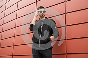 Attractive young hipster man in fashionable black clothes straightens trendy sunglasses. Stylish urban model guy stands outdoors