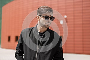 Attractive young hipster man in fashionable sunglasses in black trendy jacket with stylish hairstyle walks around the city