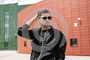 Attractive young hipster man in fashionable sunglasses in black trendy jacket with stylish hairstyle walks around the city