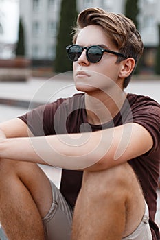 Attractive young hipster man in light shorts in stylish sunglasses with a stylish hairstyle in a trendy T-shirt sits