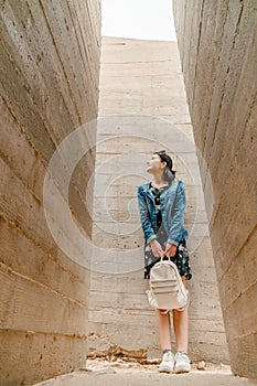 Attractive young woman stands between concrete walls. Caucasian girl in casual jeans and flower dress looks up.