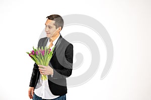 Attractive young man in black blaser holds bouquet of purple tulips in his hand and looking at left side isolated over