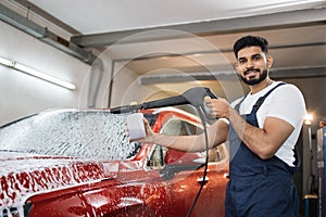 Attractive young man, car wash worker is spraying cleaning foam to a modern red luxury car