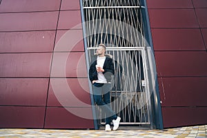 Attractive young man in casual clothers with coffee in his hands standing on office building background.