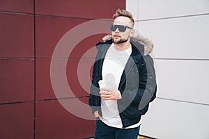 Attractive young man in casual clothers with coffee in his hands standing on office building background.