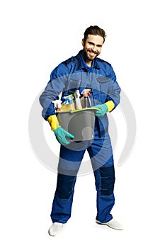 Attractive young man in cleaning uniform holding a bucket of cleaning products in his hands, isolated on white background