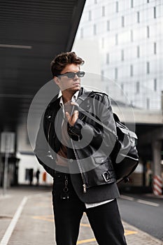 Attractive young man in dark sunglasses in a fashionable leather black jacket in retro style with a stylish backpack posing