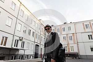Attractive young man in dark sunglasses in a fashionable leather black jacket in retro style with a stylish backpack posing