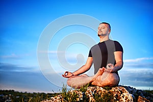 Attractive young man doing yoga on the rock against blue sky with clouds.