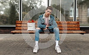 Attractive young man in fashionable blue youth jeans clothes in white stylish sneakers with vintage fabric bag sits on wooden