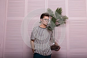 Attractive young man with a fashionable hairstyle in stylish youth summer clothes in vintage glasses is resting with a green plant