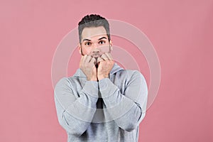 Attractive young man in gray hoodie looks scared in the camera and presses his fists to his mouth in a sign of fear and fright.