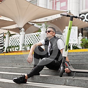 Attractive young man hipster in trendy black jeans clothes in sunglasses sits next to an electric scooter on vintage stone steps