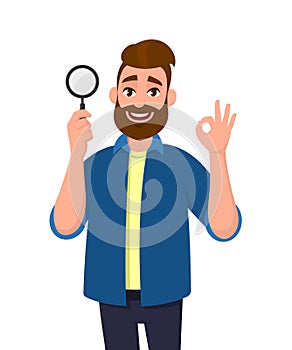 Attractive young man holding magnifying glass and gesturing okay/OK sign. Deal, good, agree, approve, search, find, discovery.