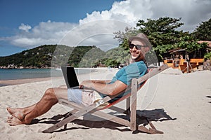 Attractive young man with laptop working on the beach. Freedom, remote work, freelancer, technology, internet, travel and vacation