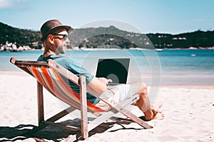 Attractive young man with laptop working on the beach. Freedom, remote work, freelancer, technology, internet, travel and vacation